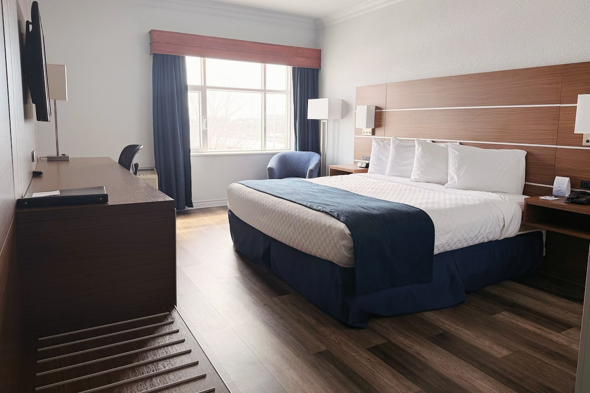 A hotel room with a bed, desk, chair, bedside table, lamp, and a window with curtains.