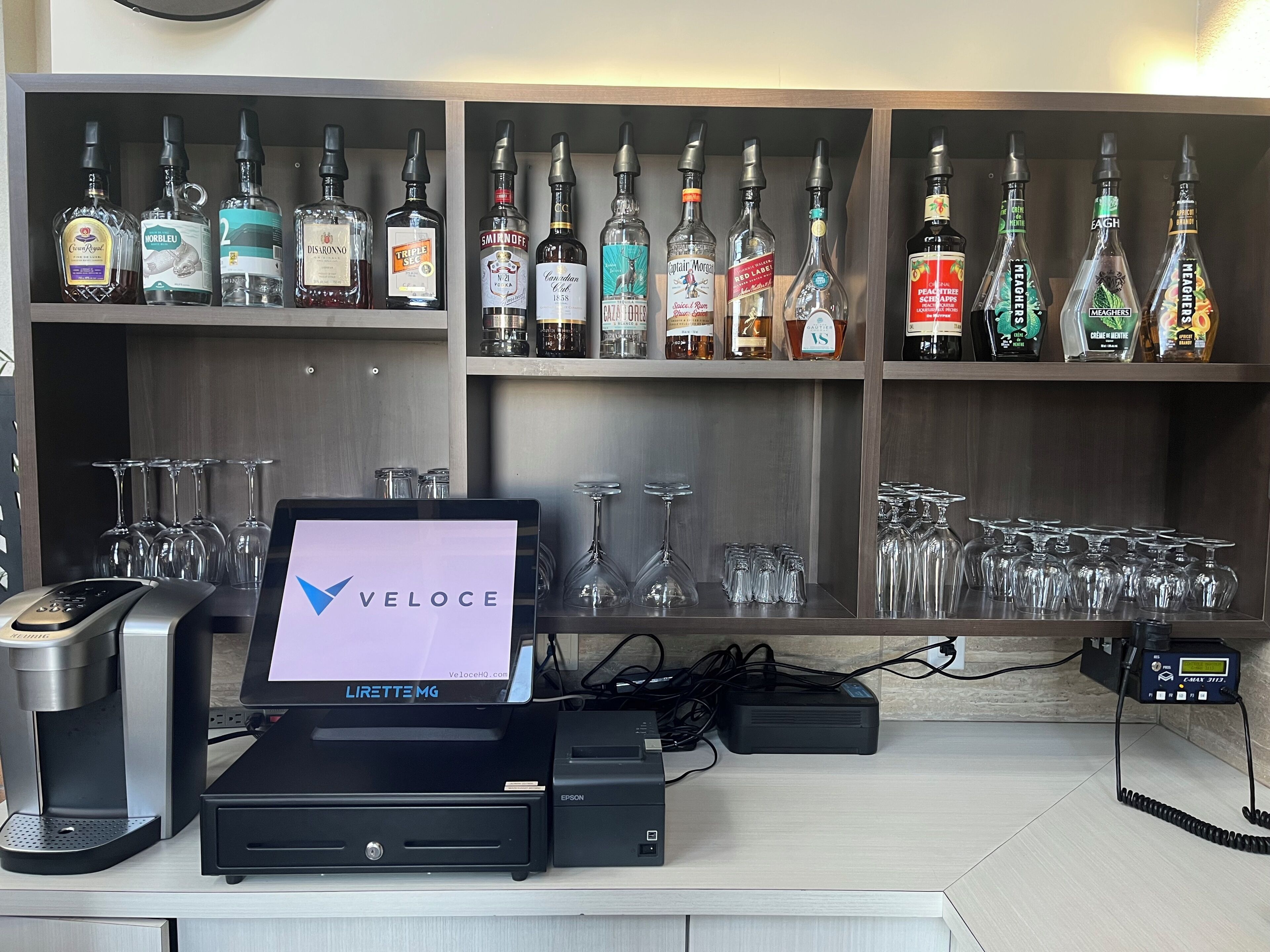 A bar area with a Veloce digital screen, a coffee machine, and a variety of liquor bottles.