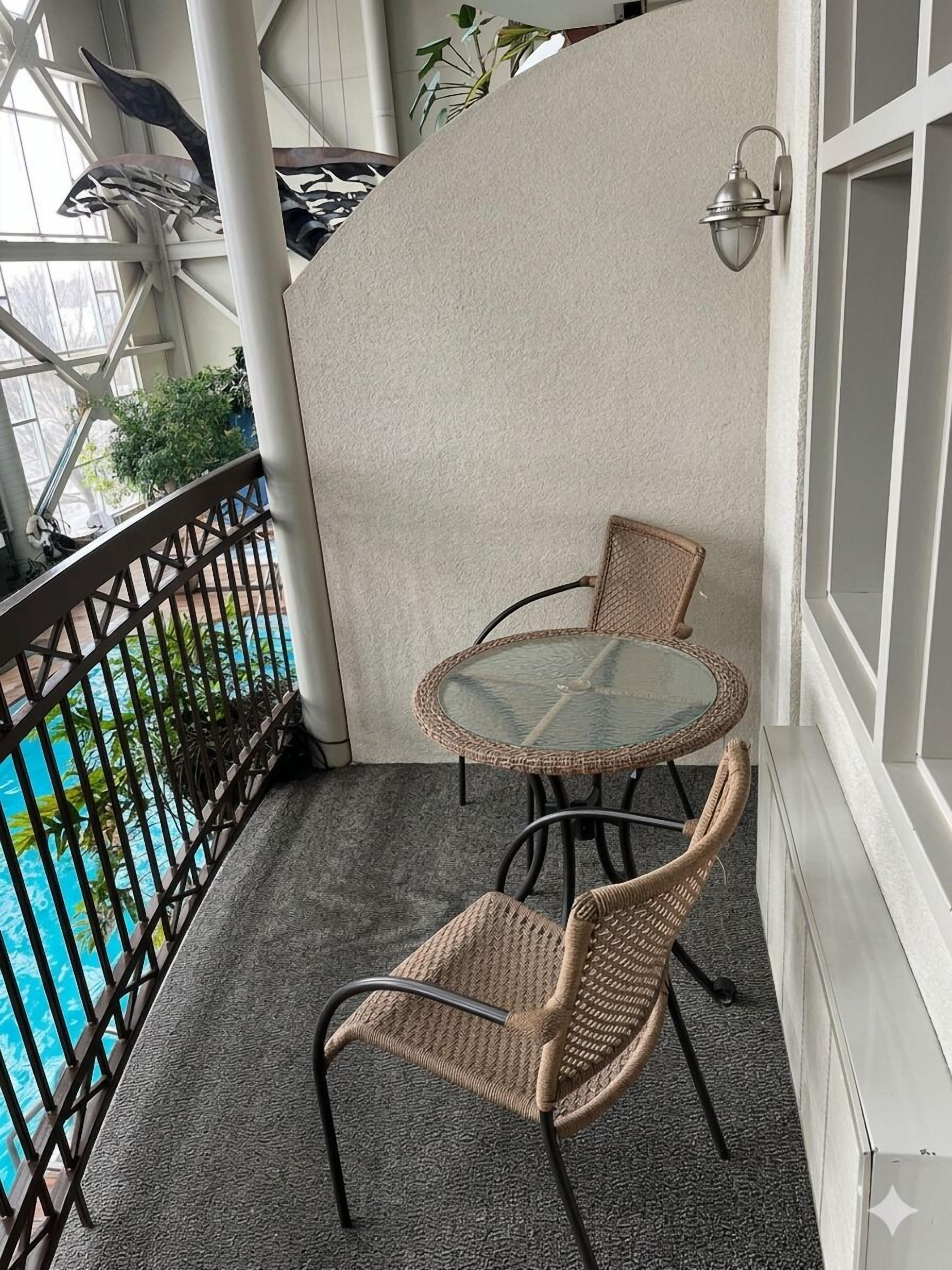 Un balcó amb taula de vidre i cadires de vimetla, una piscina al fons i una paret decorativa.