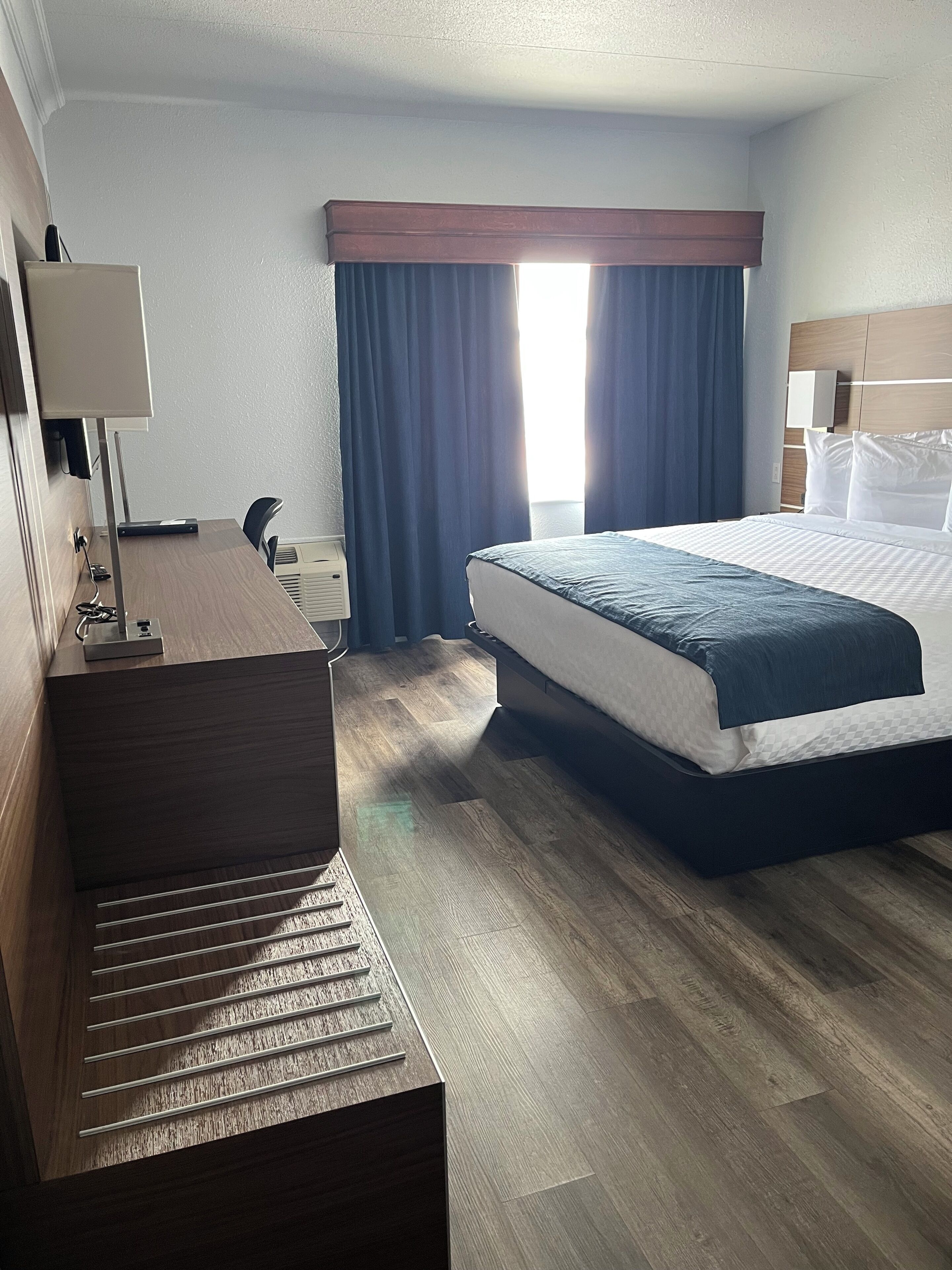 A hotel room with a bed, a desk with a lamp, a chair, and a window with curtains.