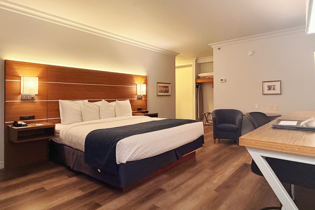 A hotel room with a wooden headboard, a bed with white and navy bedding, a desk with a chair, and a small table with a lamp.