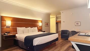 A hotel room with a wooden headboard, a bed with white and navy bedding, a desk with a chair, and a small table with a lamp.