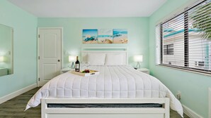 A bedroom with a bed, a nightstand, a window with blinds, a door, and a wall with a painting.