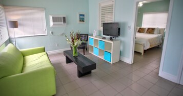 A modern living room with a green sofa, a black coffee table with a vase, a TV on a white stand, and a bed with a blue and white cabinet.