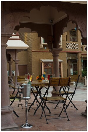 An outdoor dining area with a table set for two, surrounded by ornate columns and a tiled floor.