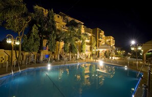 A nighttime pool area with a clear pool, surrounding palm trees, and a building with balconies. - Niralidhani Ethnic Resort (Jodhpur)