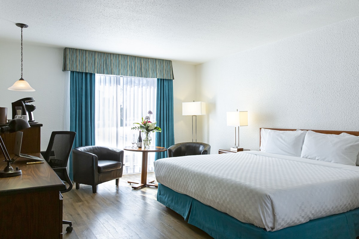 A hotel room with a bed, two chairs, a desk with a lamp, a small table with a vase, and a window with curtains.