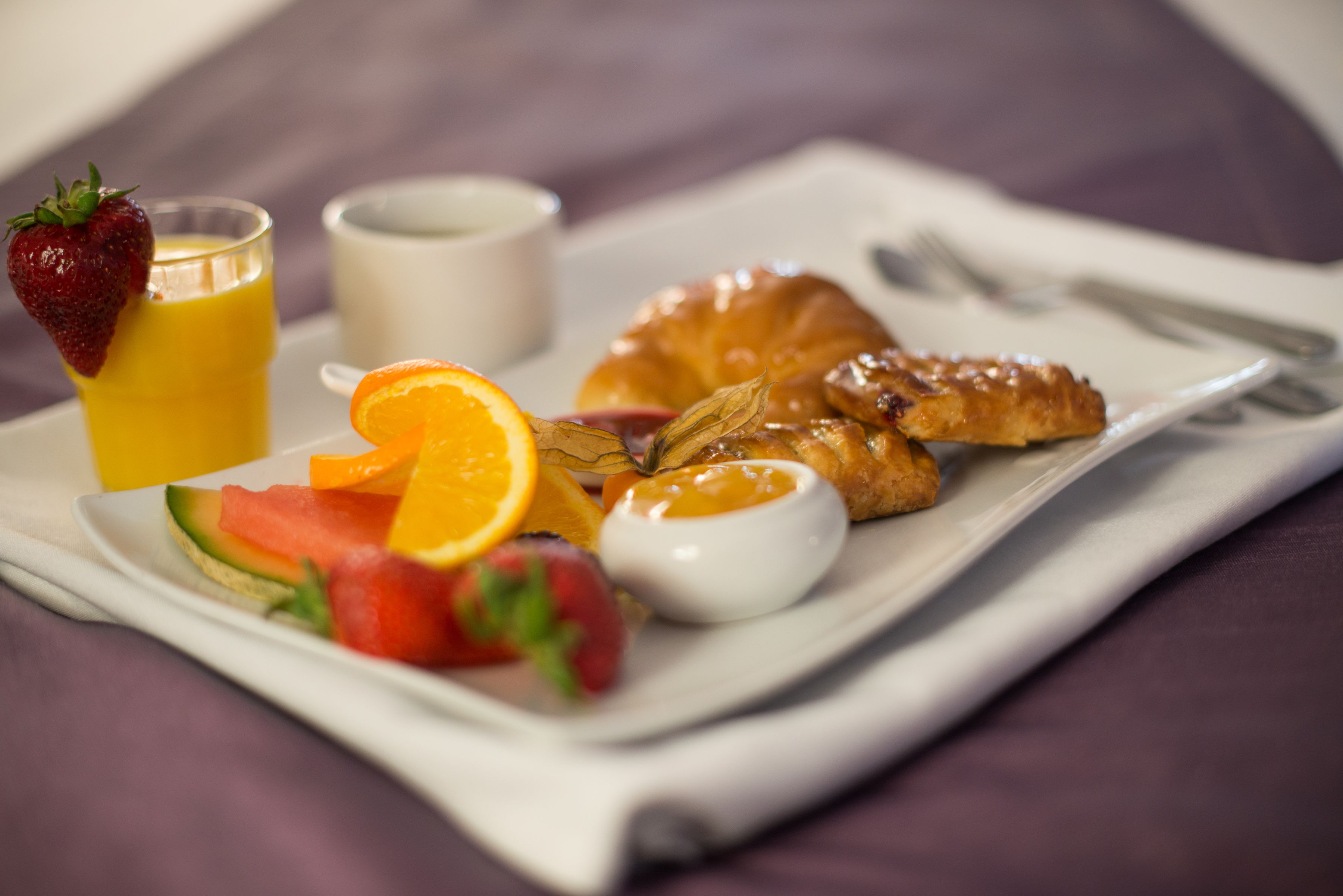 A breakfast tray with a croissant, strawberries, watermelon, orange slices, a glass of orange juice, and a small bowl of yogurt.