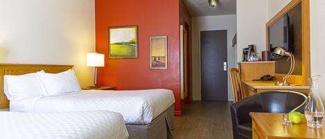 A hotel room with two beds, a desk with a lamp, a chair, and a small table with a vase and an apple.