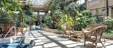 An indoor pool area with a glass ceiling, surrounded by various plants and wicker furniture.