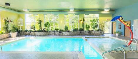 An indoor swimming pool with clear blue water, surrounded by potted plants and a play area with a slide.