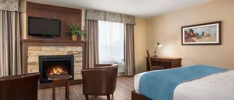 A hotel room with a fireplace, a bed with a blue blanket, a television, and a painting on the wall.