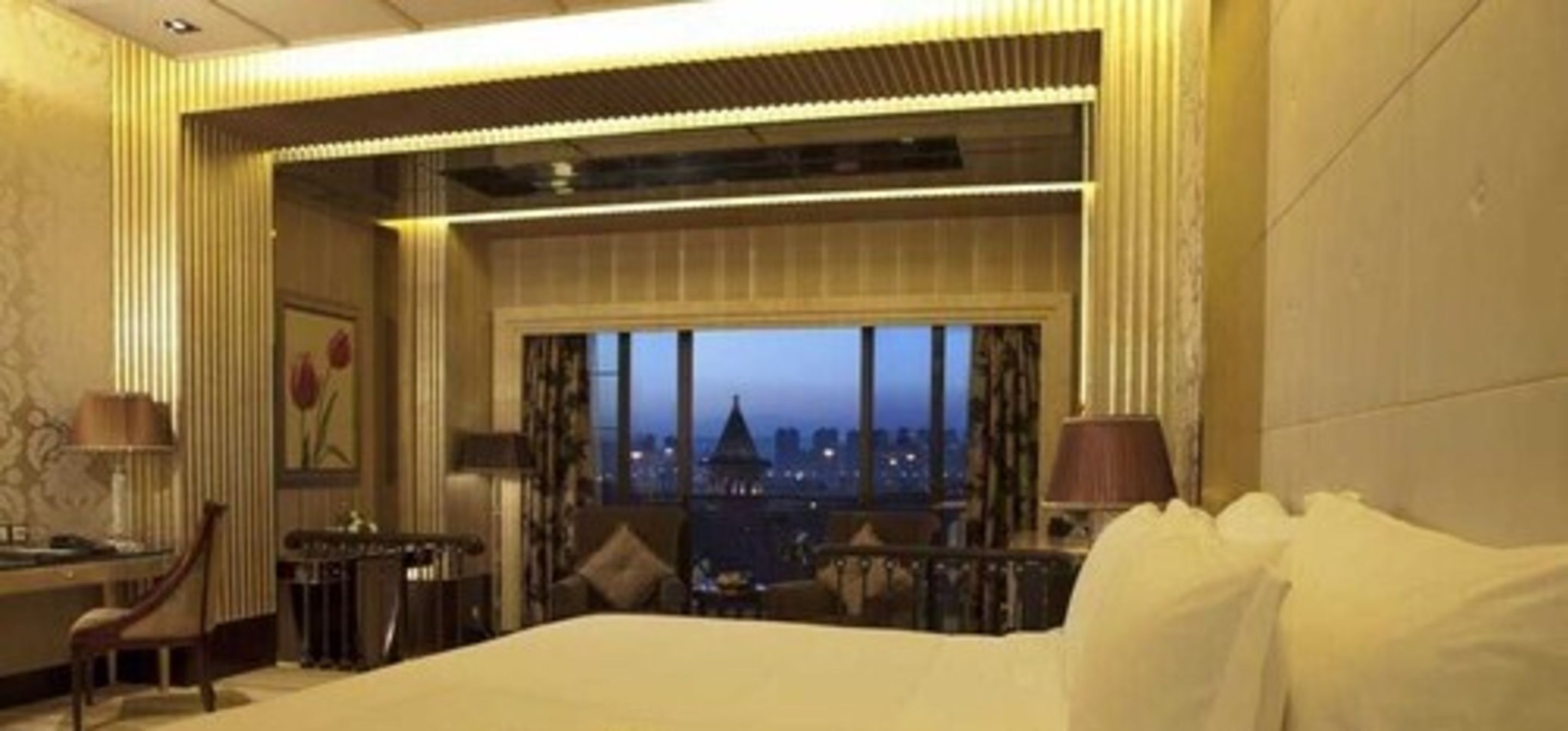 A hotel room with a large bed, a desk with a chair, a lamp, and a view of a cityscape at dusk.