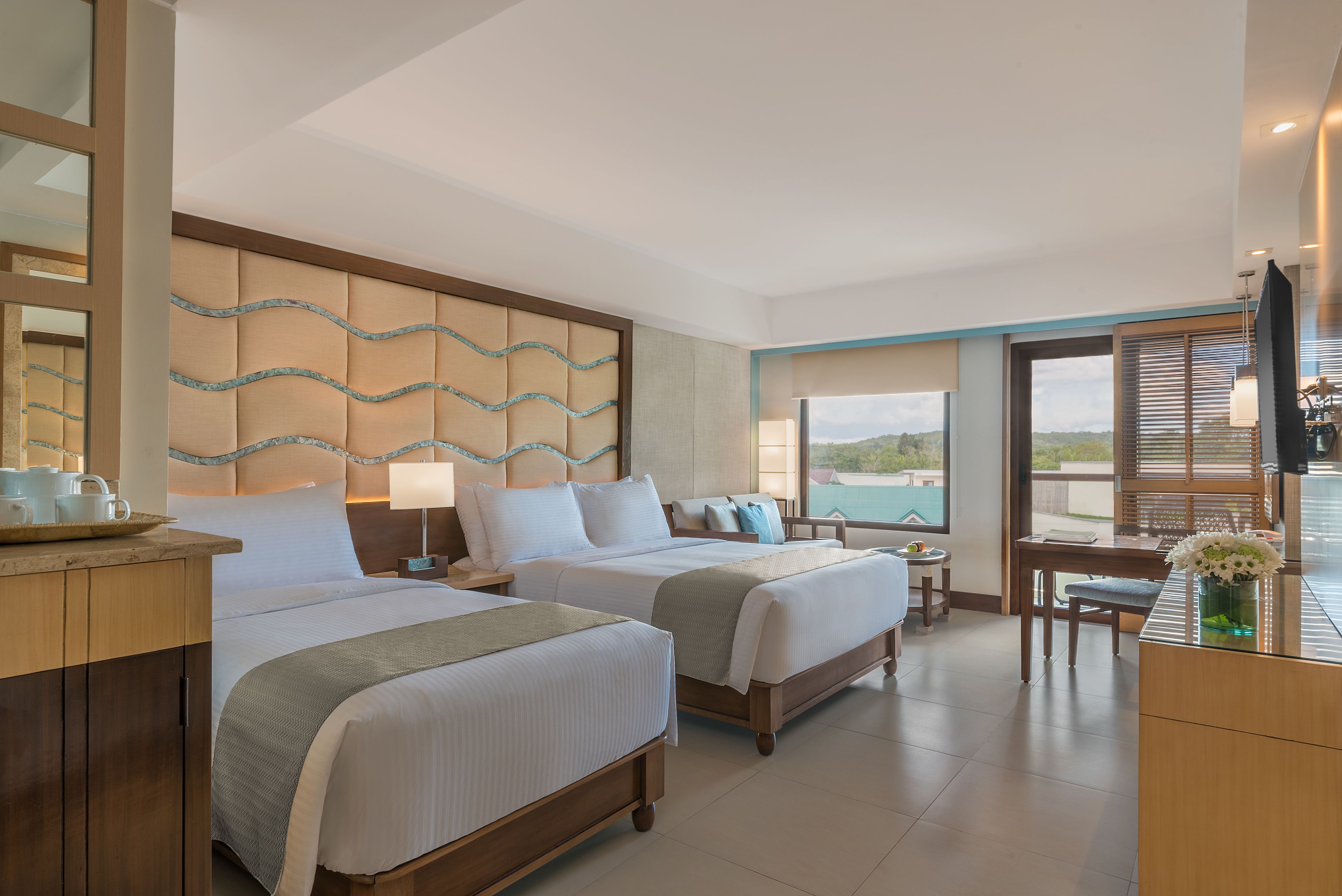 A hotel room with two beds, a desk, a chair, a TV, and a view of the outdoors.