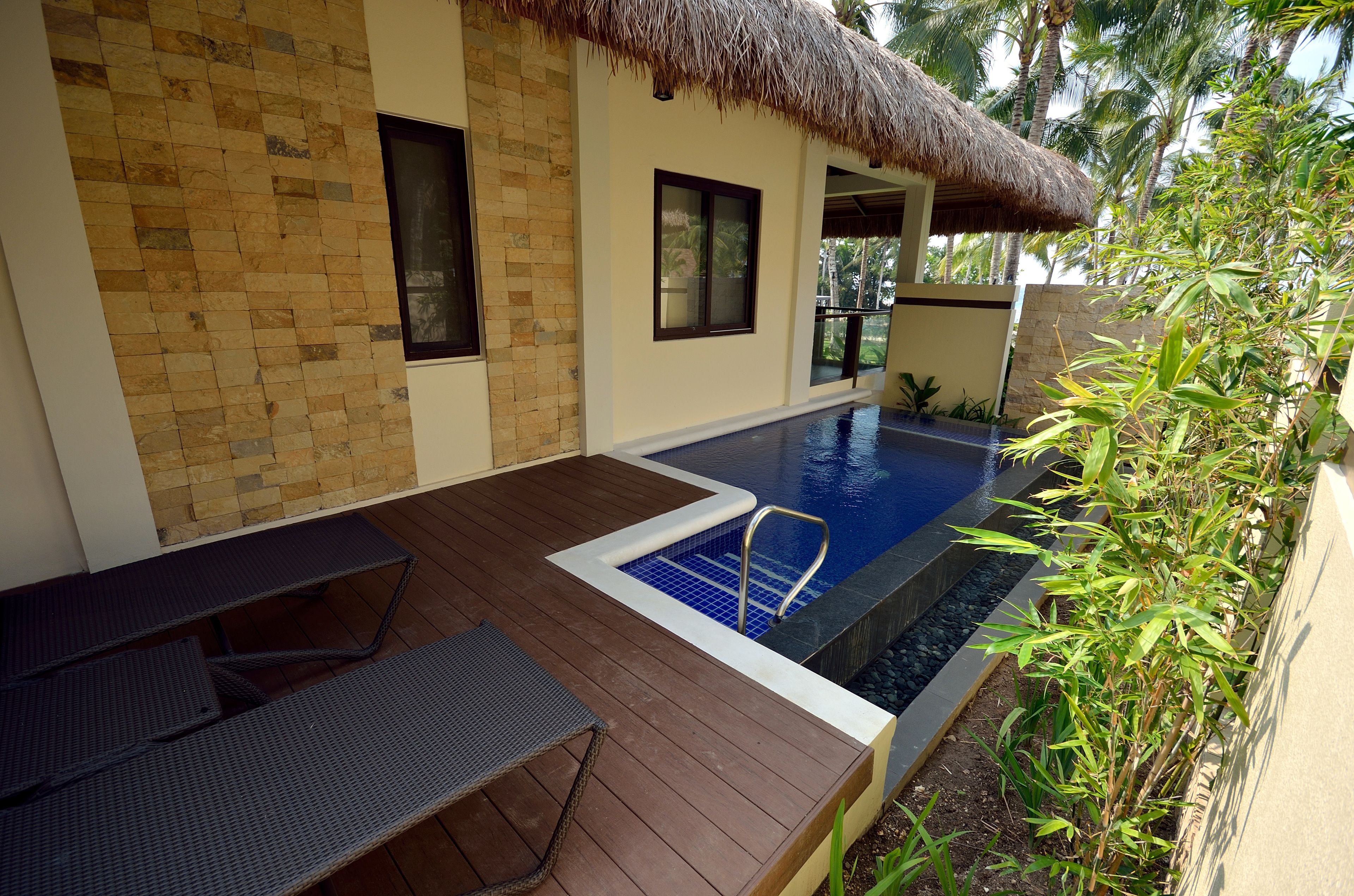 A pool area with a wooden deck, a thatched roof structure, and a view of palm trees.