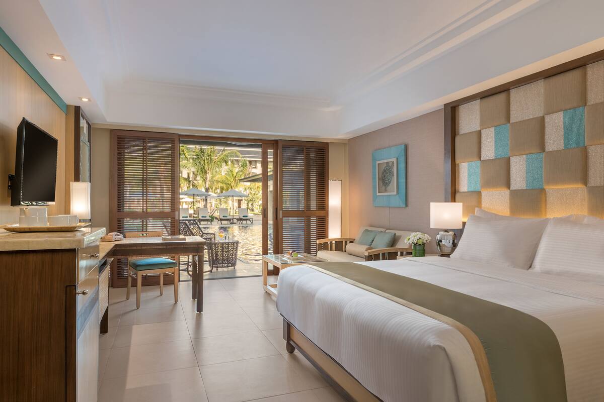 A hotel room with a large bed, a desk with a chair, a television, and a view of the outdoor area.