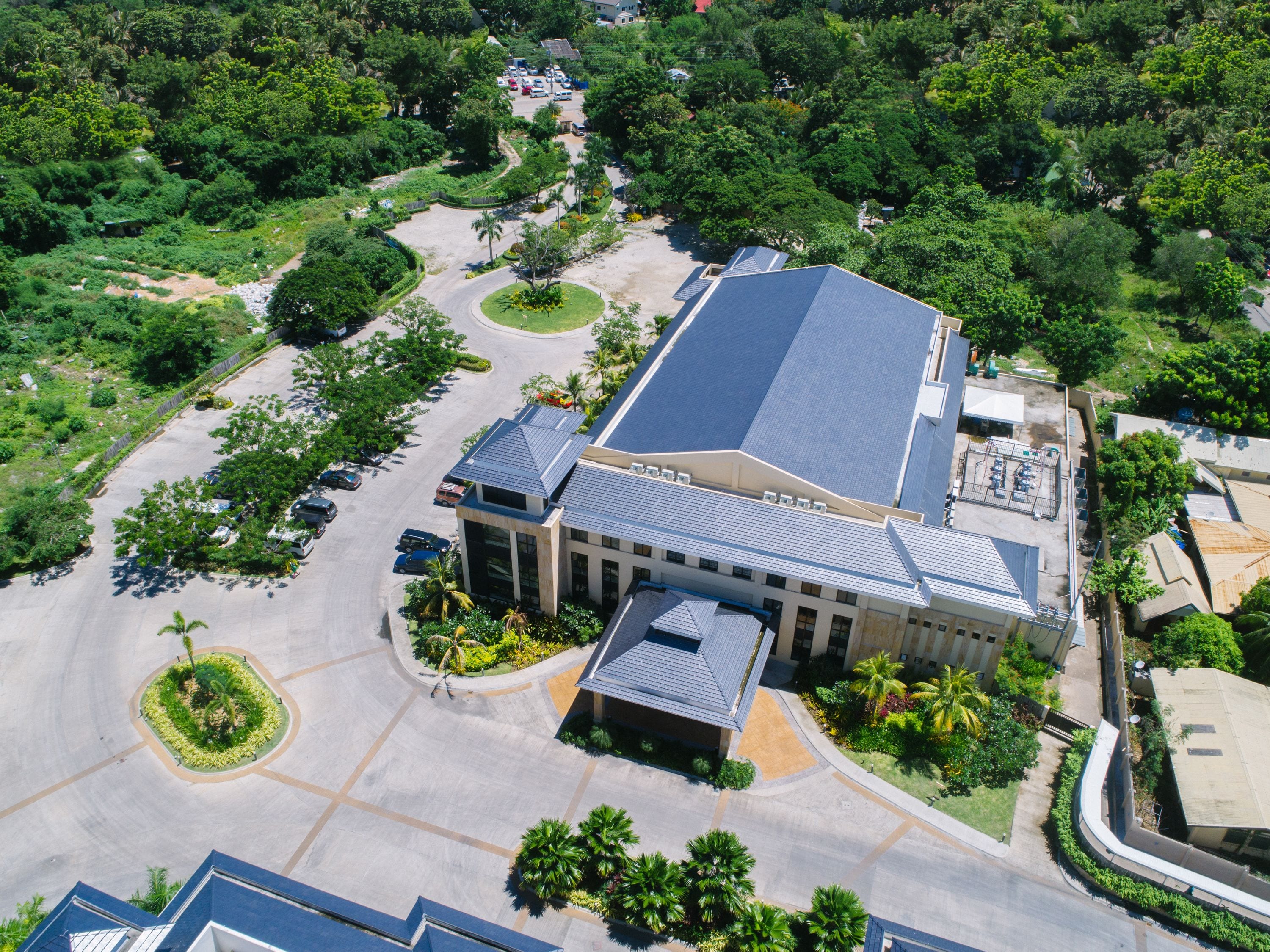 An aerial view of a building with a flat roof, surrounded by greenery and a parking lot.