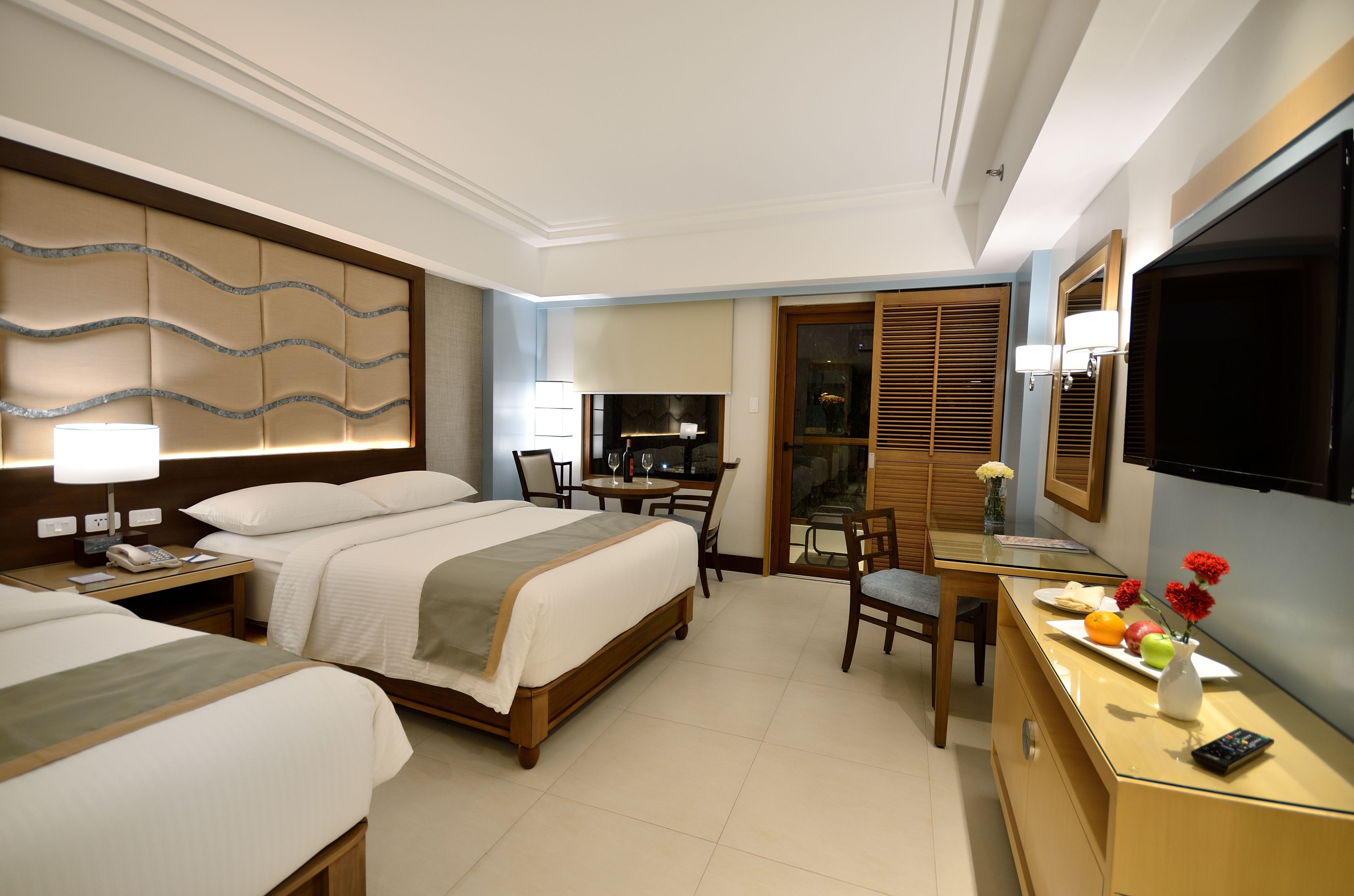 A hotel room with two beds, a desk with a chair, a TV, and a dining area with a table and chairs.