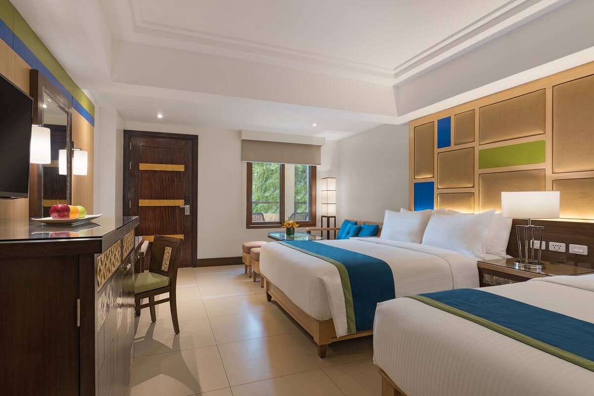 A hotel room with a large bed, a smaller bed, a desk with a chair, a TV, and a window with a view of trees.