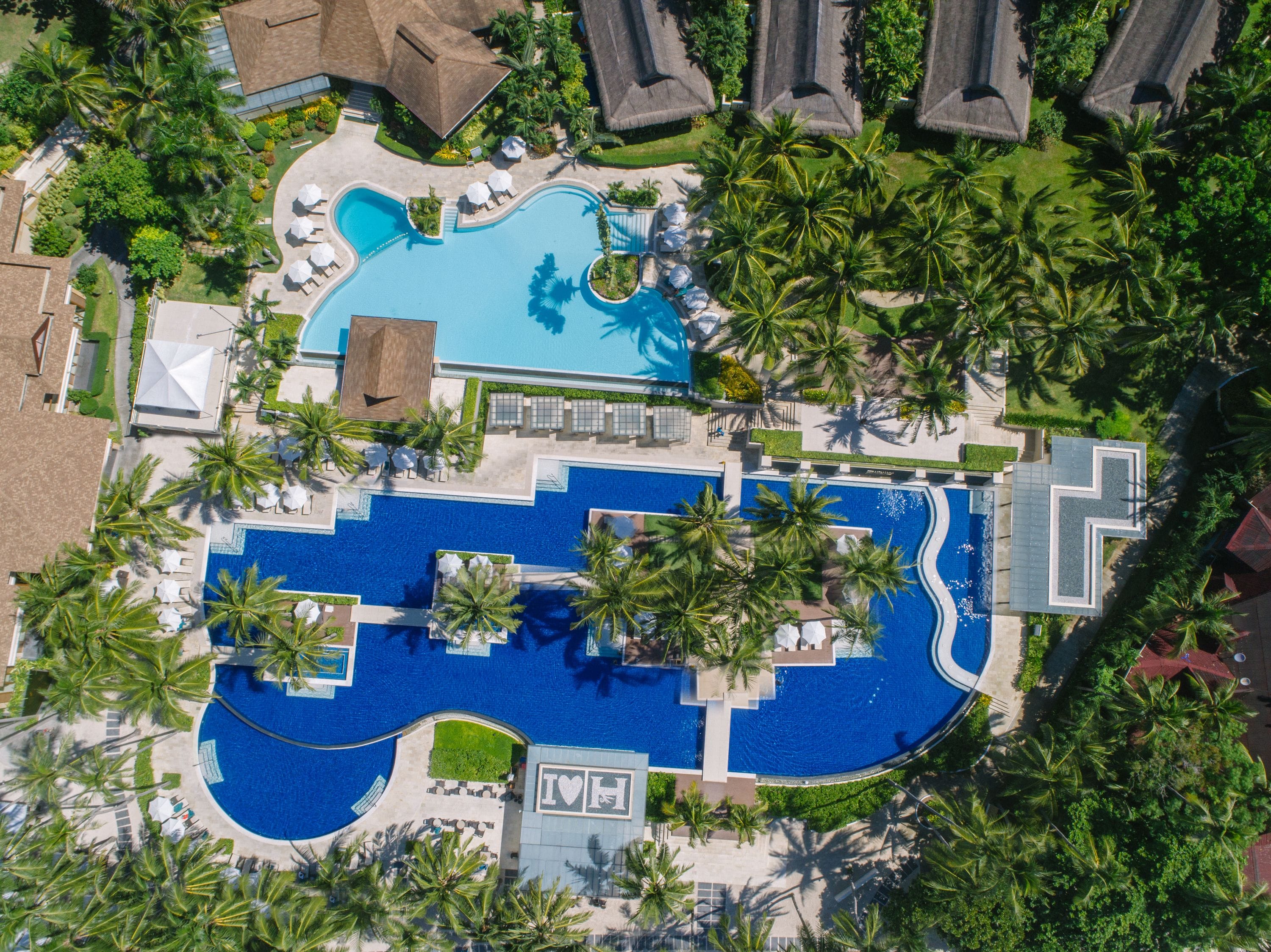 Eine Luftaufnahme eines Resorts mit einem großen zentralen Pool, mehreren kleineren Pools und zahlreichen Palmen.