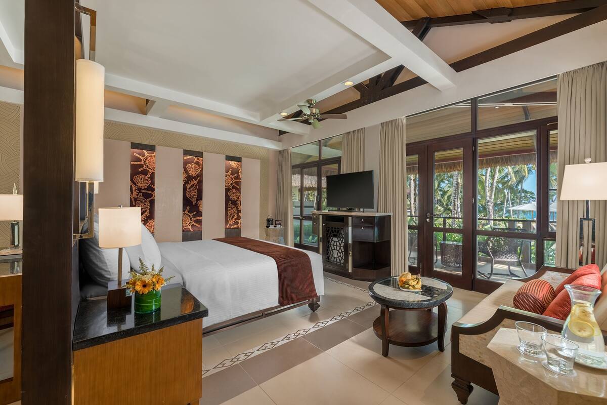 A hotel room with a bed, a desk, a TV, a dining area, and large windows with a view of palm trees.