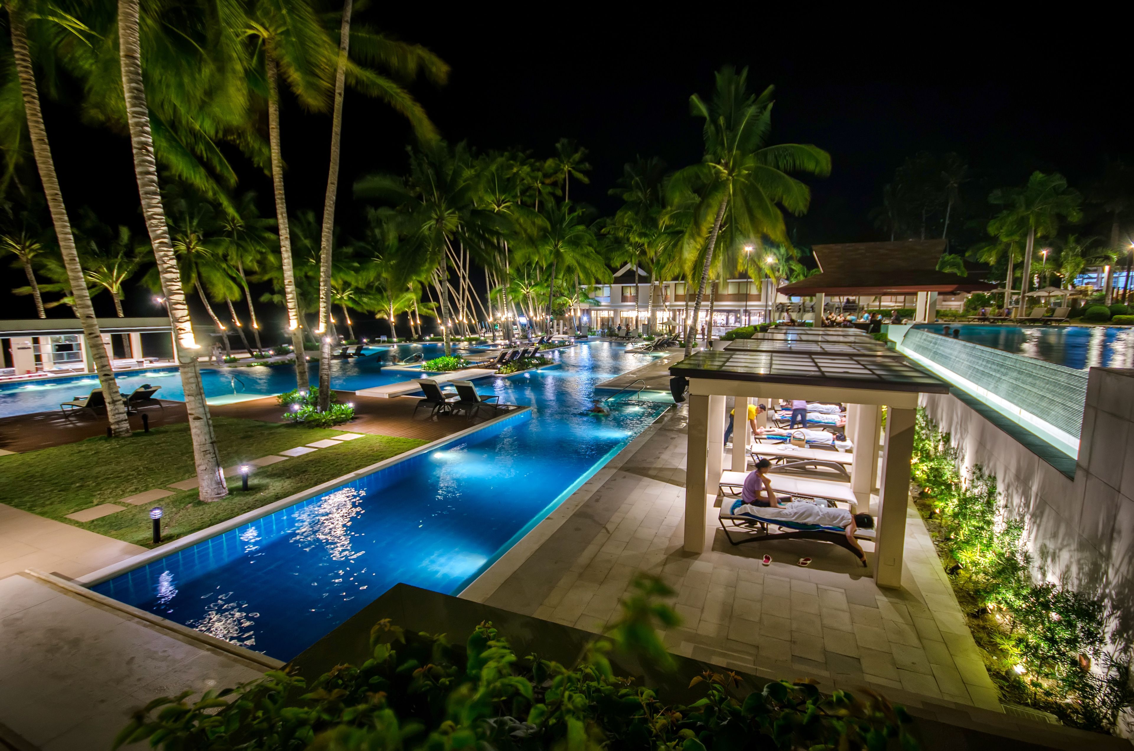 Vista noturna de uma área de piscina em um resort, com palmeiras, espreguiçadeiras e um espaço coberto para sentar.