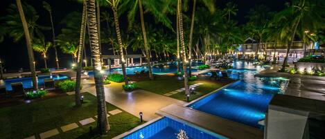 A nighttime resort with multiple swimming pools, palm trees, and well-lit pathways.