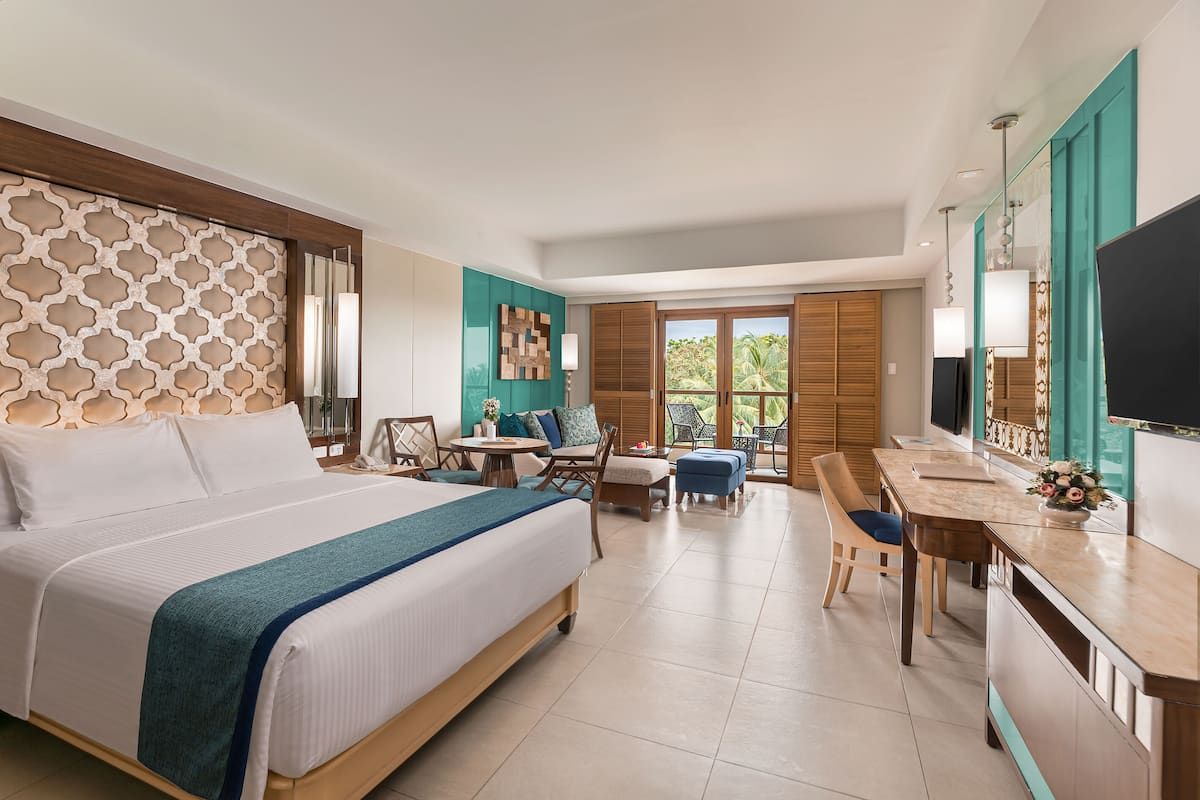 A hotel room with a large bed, a desk, a chair, a TV, and a balcony with a view of palm trees.