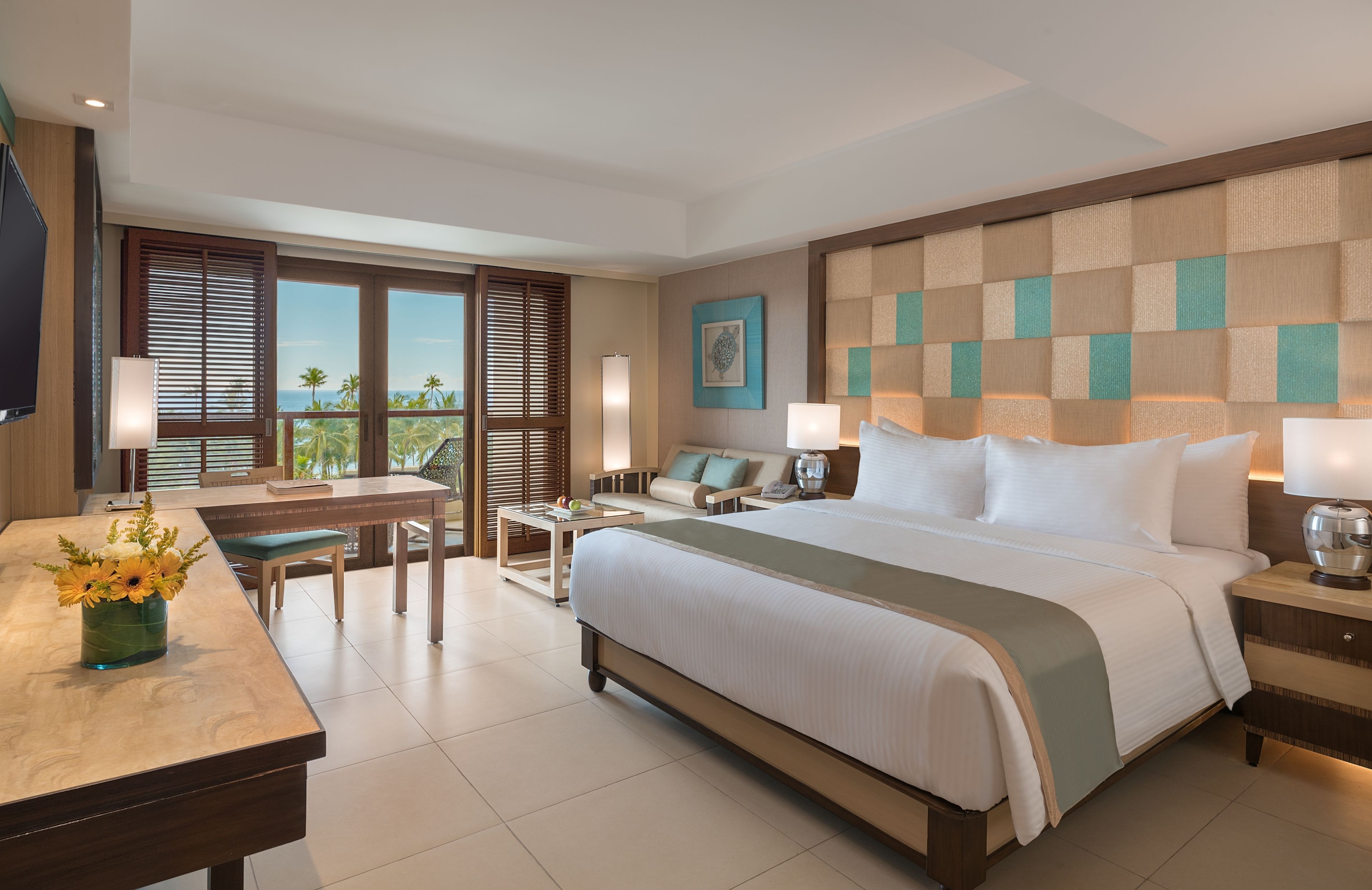 Habitación de hotel con una cama grande, un escritorio, una silla, un televisor y vista a palmeras.