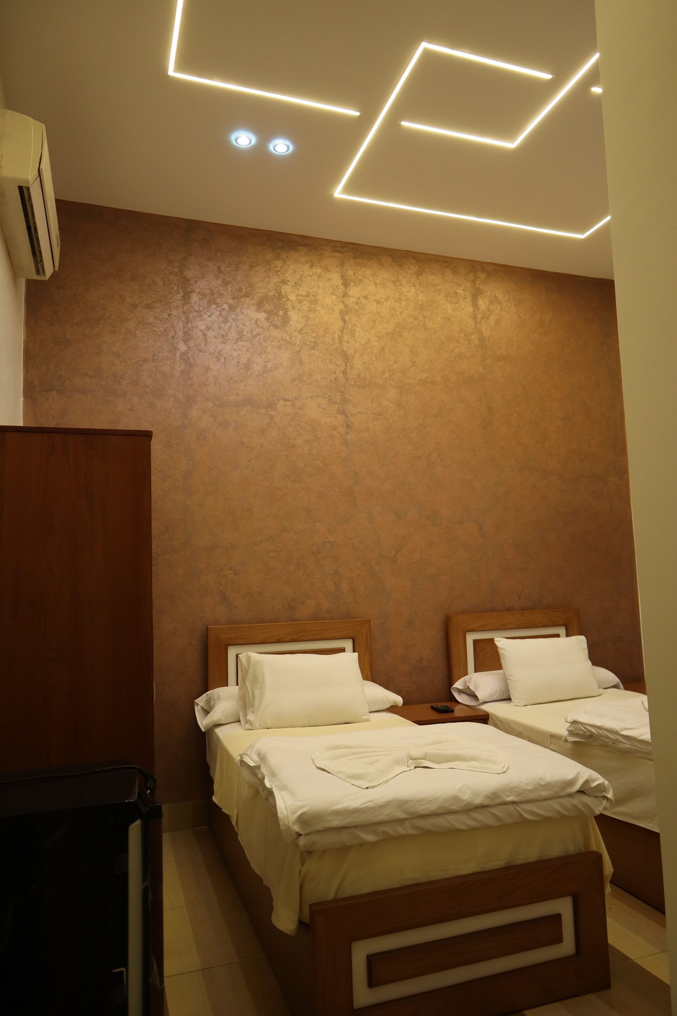 Une chambre d’hôtel avec deux lits, une armoire en bois et un plafond orné de luminaires aux motifs géométriques.