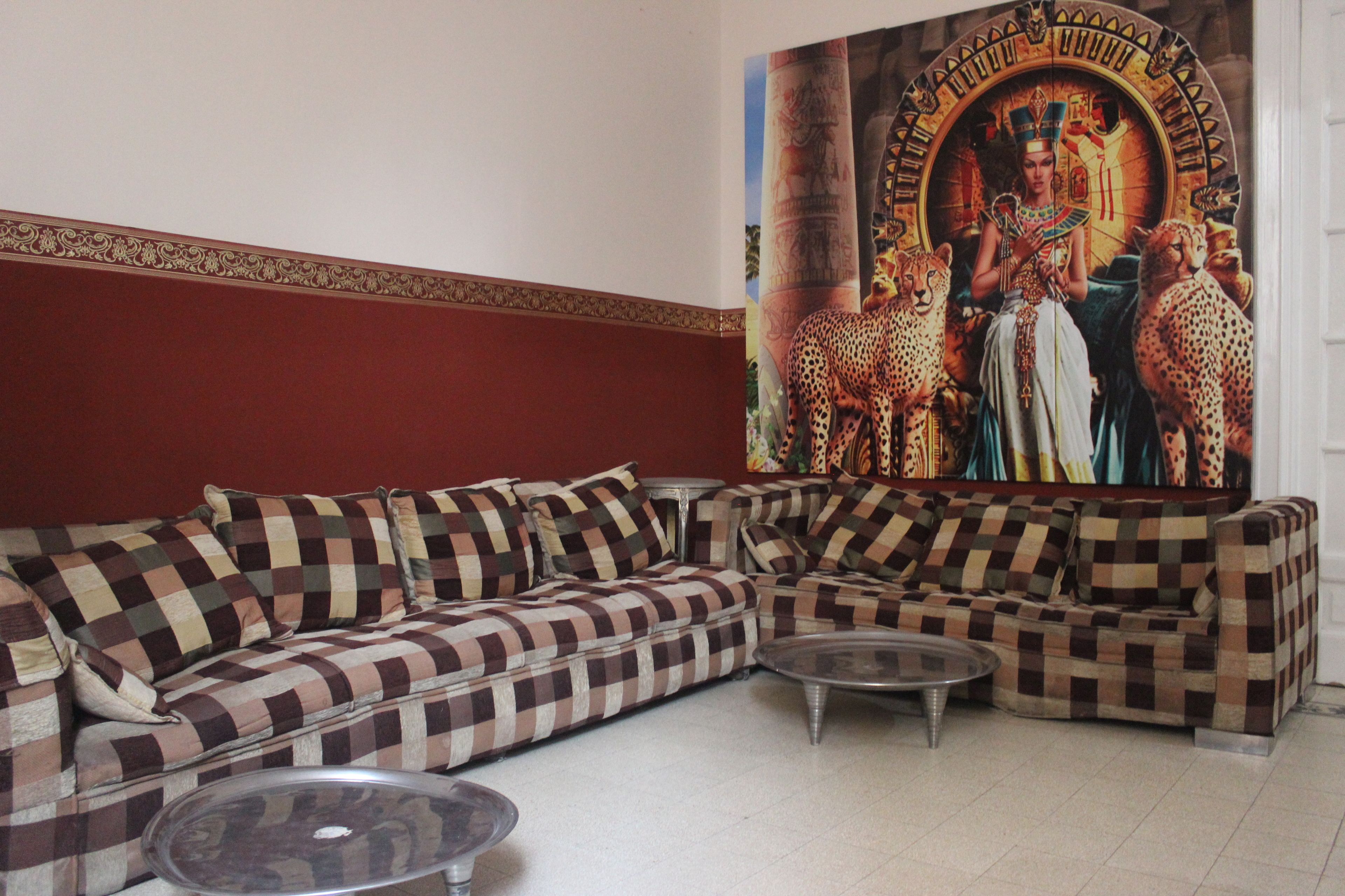 A living room with a checkered sofa, two glass coffee tables, and a large wall mural depicting ancient Egyptian figures and animals.
