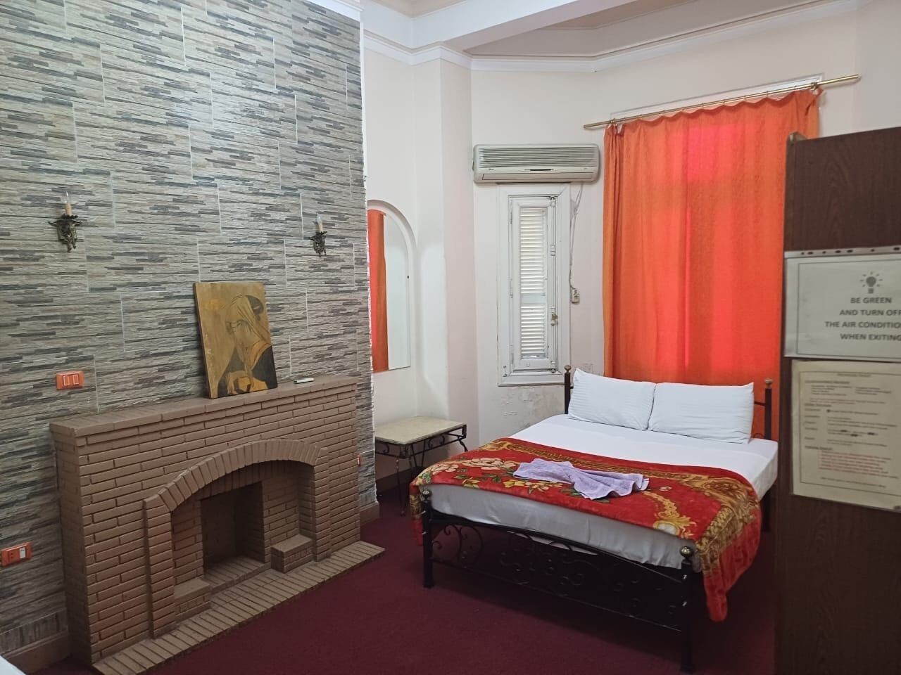 Une chambre avec une cheminée en briques, un lit recouvert d’une courtepointe rouge et or, un appareil de climatisation et une fenêtre avec des rideaux orange.