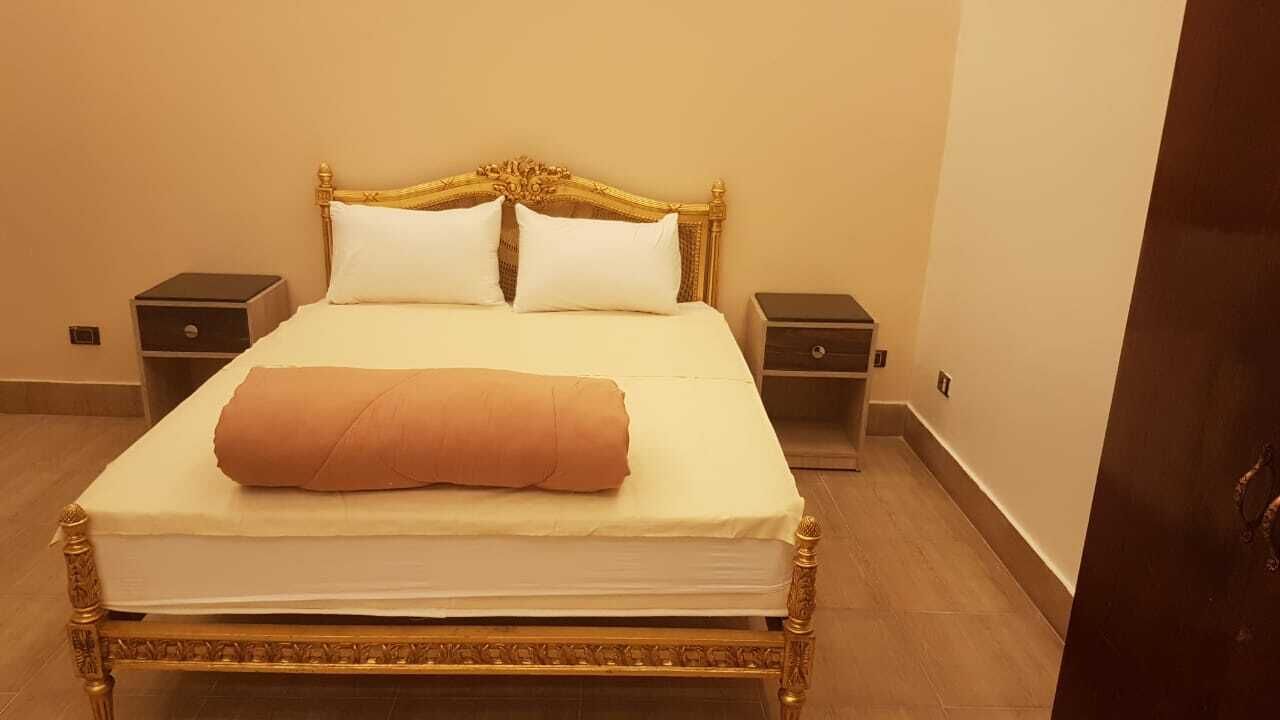 Un lit avec une tête de lit dorée, une literie blanche et une couverture marron. Il y a deux tables de chevet avec des tiroirs.