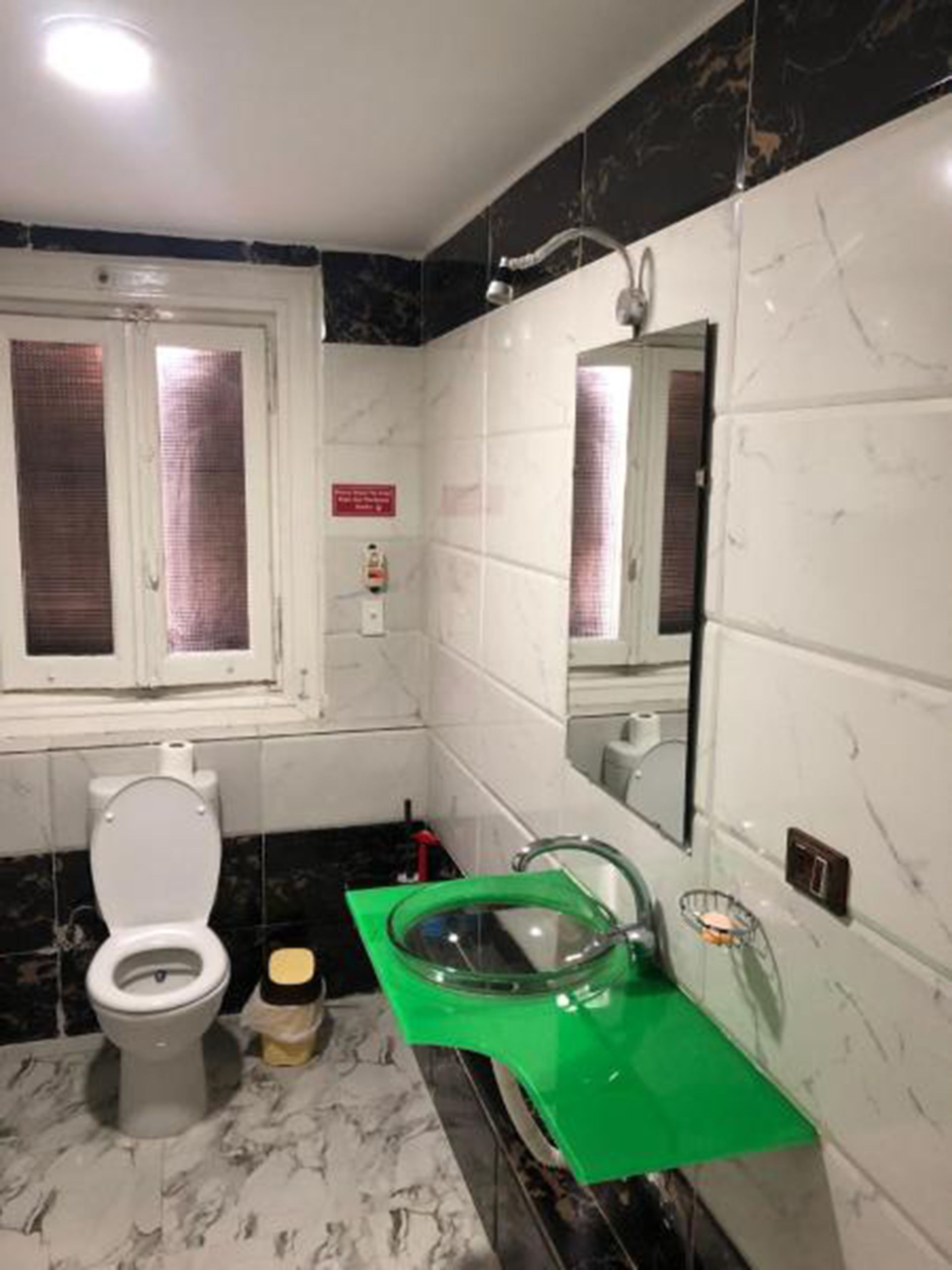 A bathroom with a toilet, a green sink, a mirror, and a window with blinds.