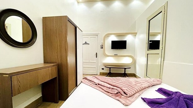 A bedroom with a bed, a wooden bench, a mirror, a TV, and a bathroom door. - Meramees Hostel (Cairo)