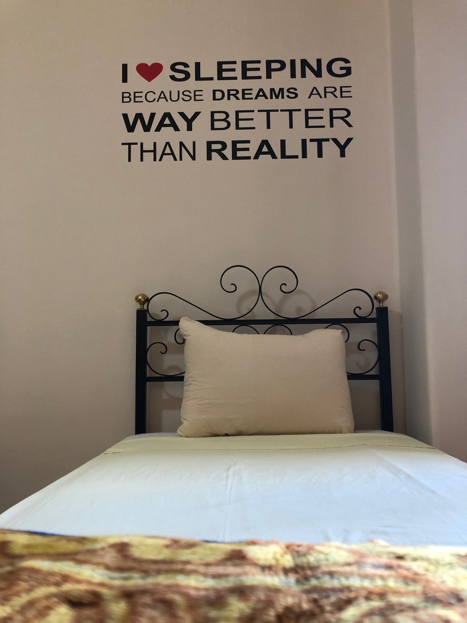 Uma cama bem arrumada com lençol branco e travesseiro bege, uma frase decorativa na parede e cabeceira de metal.