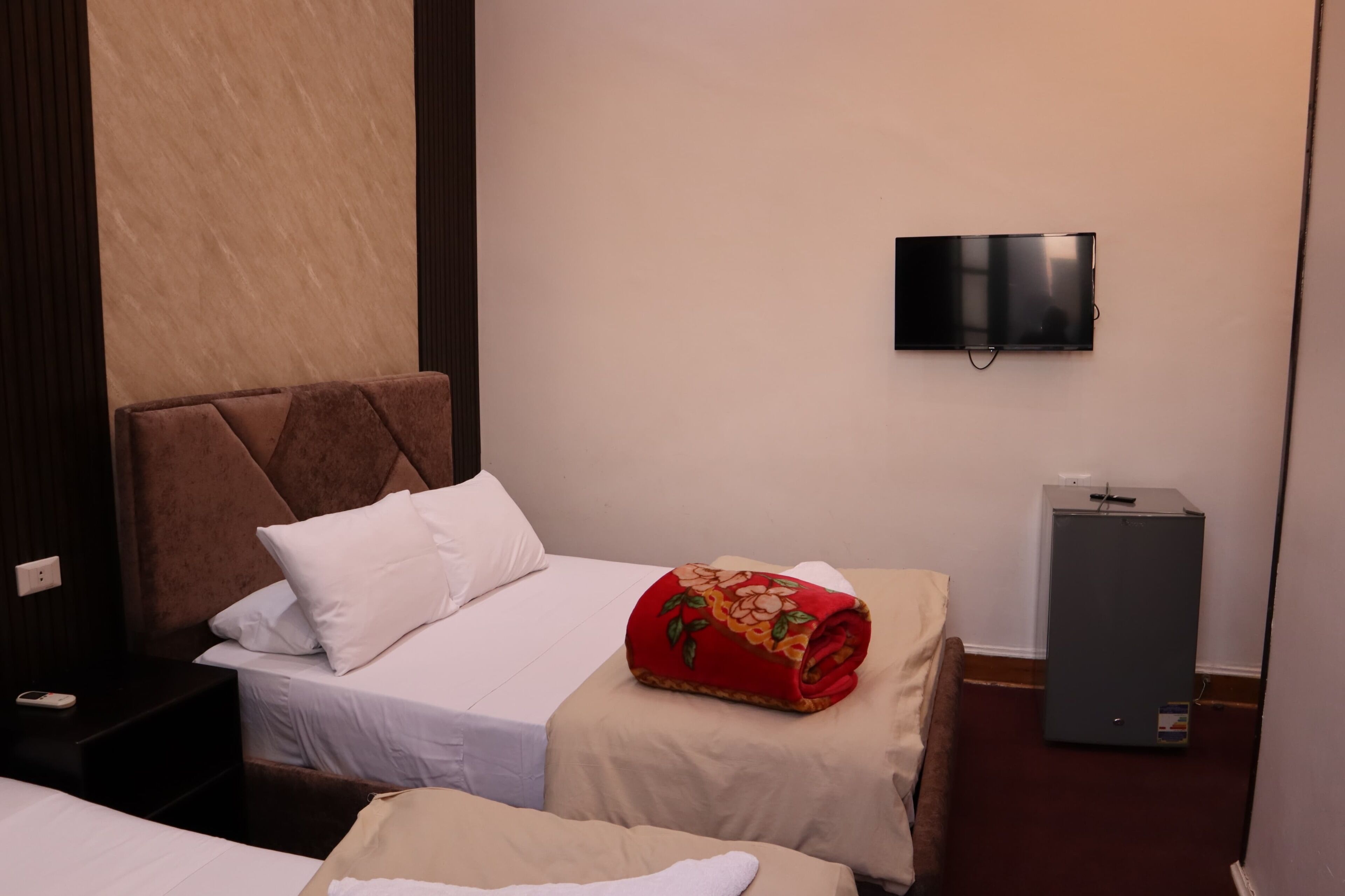 A hotel room with a bed, a television mounted on the wall, a small cabinet, and a red floral blanket on the bed.