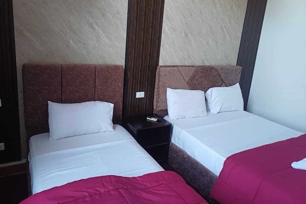 Habitación de hotel con dos camas, cada una con ropa de cama blanca y un detalle en rojo. Un mesita de noche entre las camas contiene un teléfono y una lámpara pequeña. Las paredes presentan un diseño texturizado.
