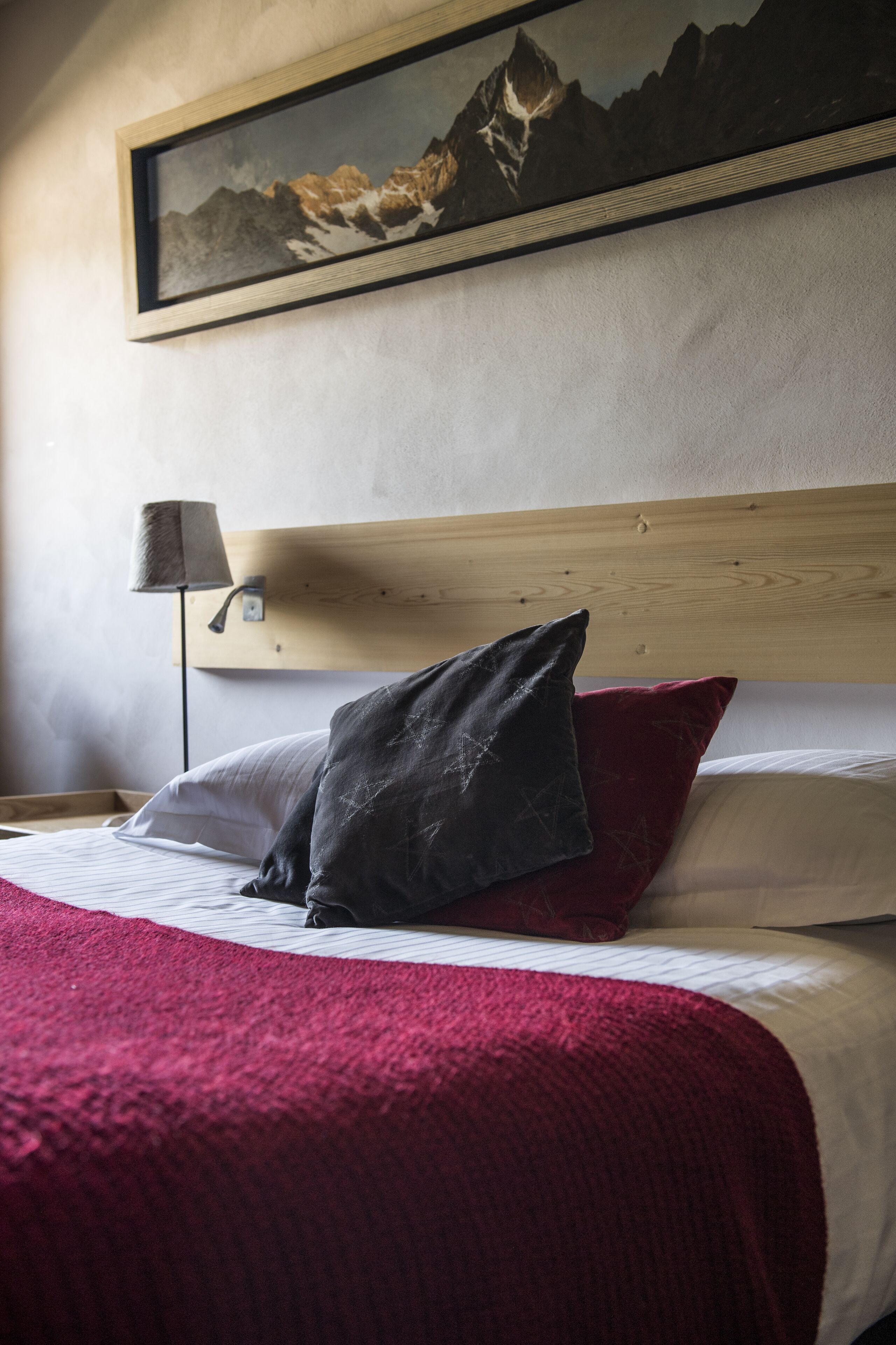 A neatly made bed with a red blanket, a wooden headboard, and a mountain painting above it.