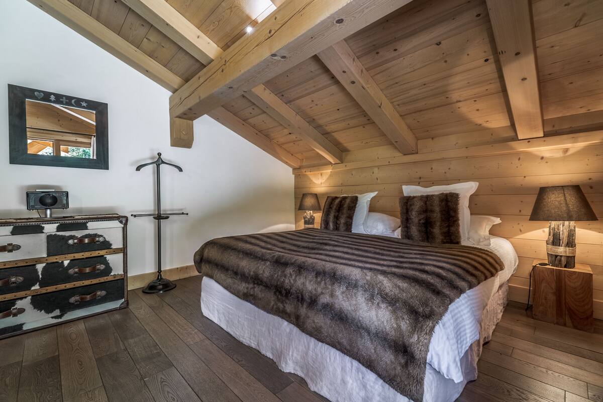 Un dormitorio con techo de madera, una cama con manta de piel, un tocador con cajones, un espejo y una lámpara.