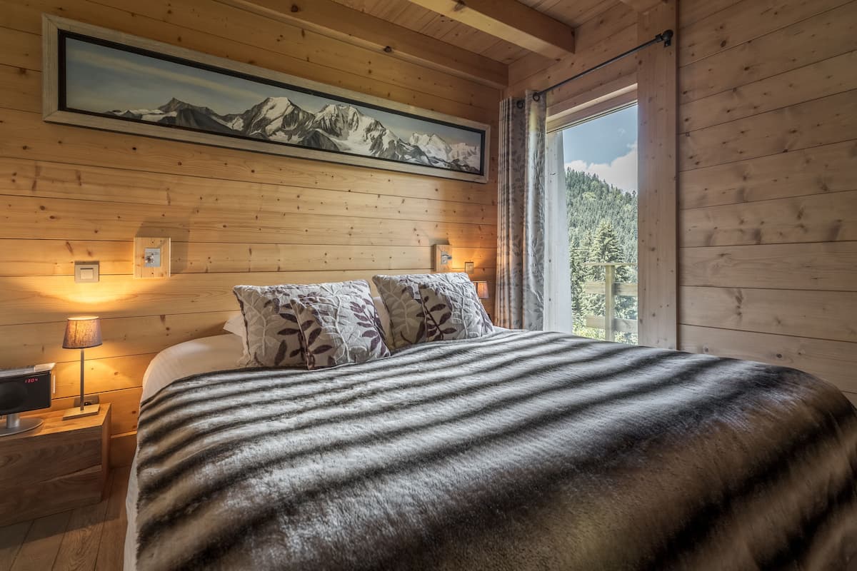 Un dormitorio de cabaña de madera con una cama, dos lámparas de noche, una mesita de noche, un cuadro de un paisaje montañoso y una ventana con vistas a árboles.