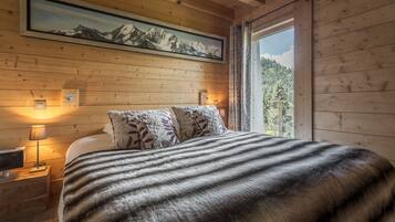 Une chambre à coucher dans une cabane en bois, avec un lit, deux lampes de chevet, une table de chevet, un tableau représentant une montagne encadré et une fenêtre donnant sur la forêt.