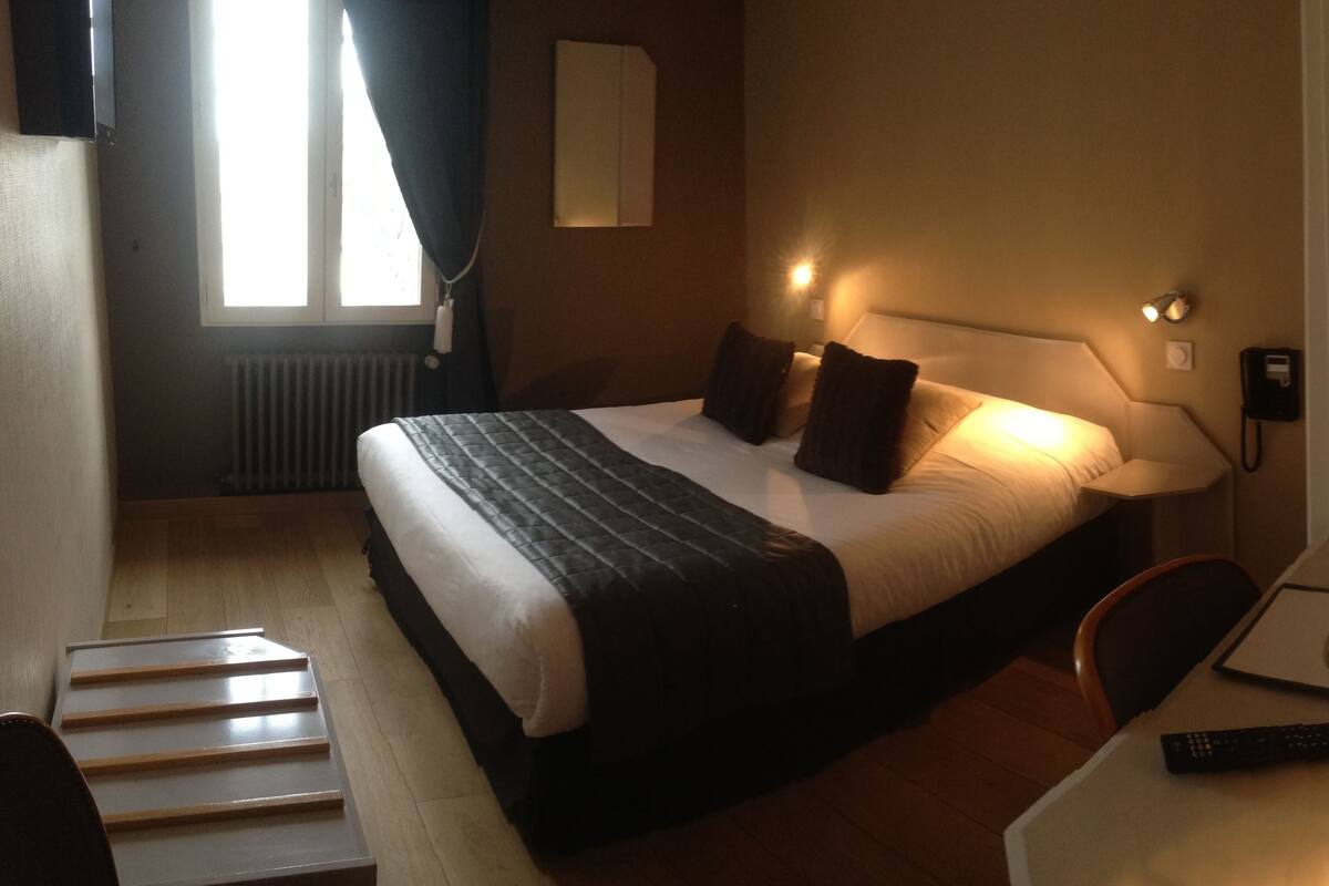 A hotel room with a bed, bedside table, lamp, and a remote control on the table.