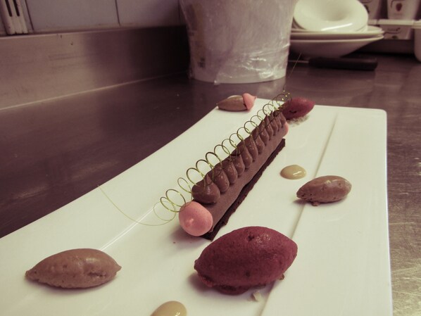 Un postre sobre un plato blanco, con detalles de chocolate y tonos rosados, colocado sobre una mesa en una cocina.