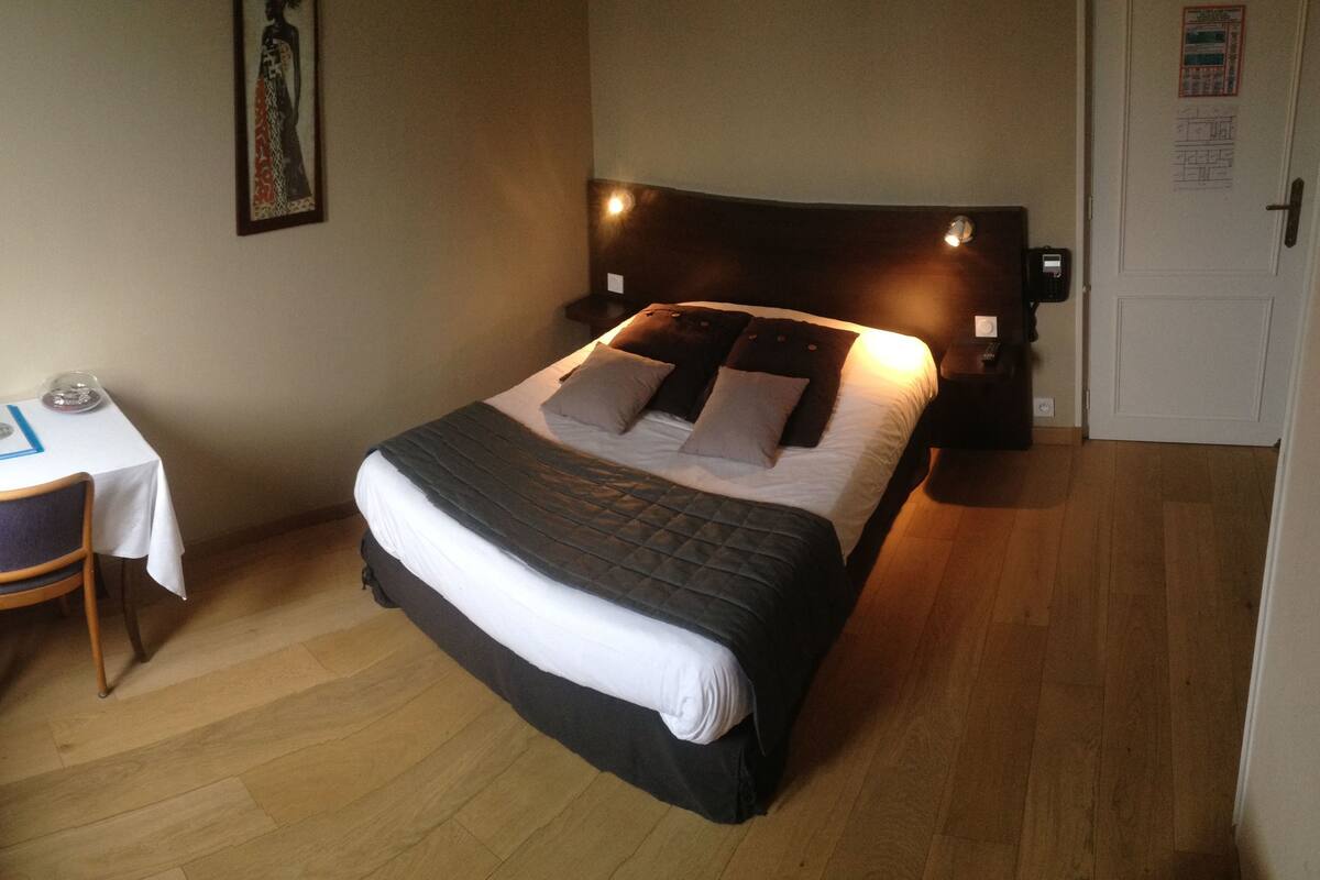 A neatly arranged bedroom with a bed, bedside table, chair, and a small desk with a lamp and book.