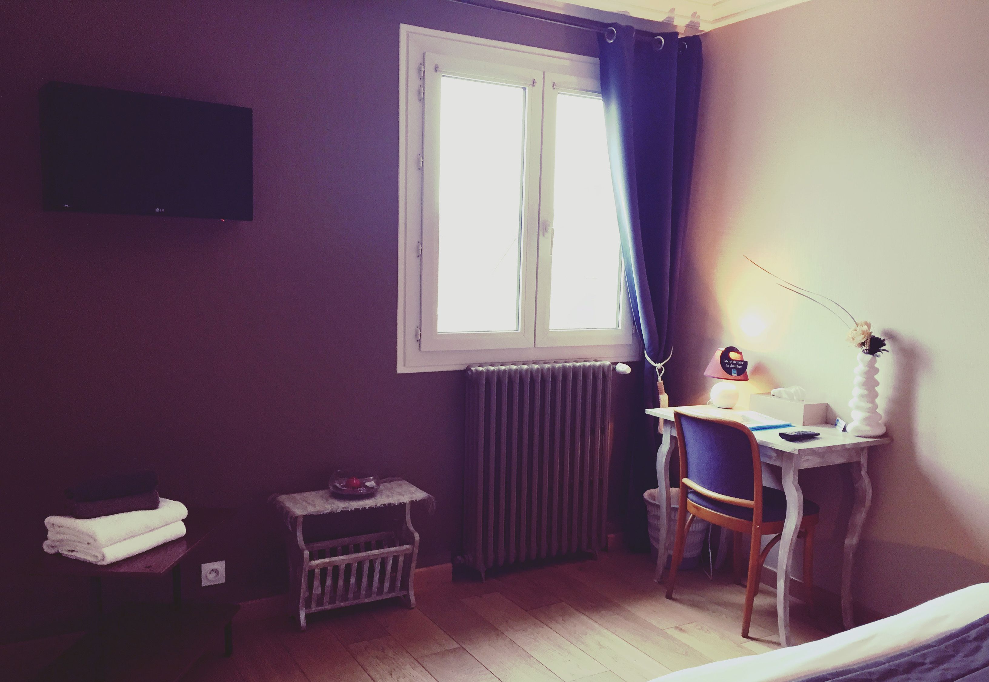 Una habitación con una cama, un escritorio con una silla, un radiador, una ventana con cortinas y un televisor montado en la pared.
