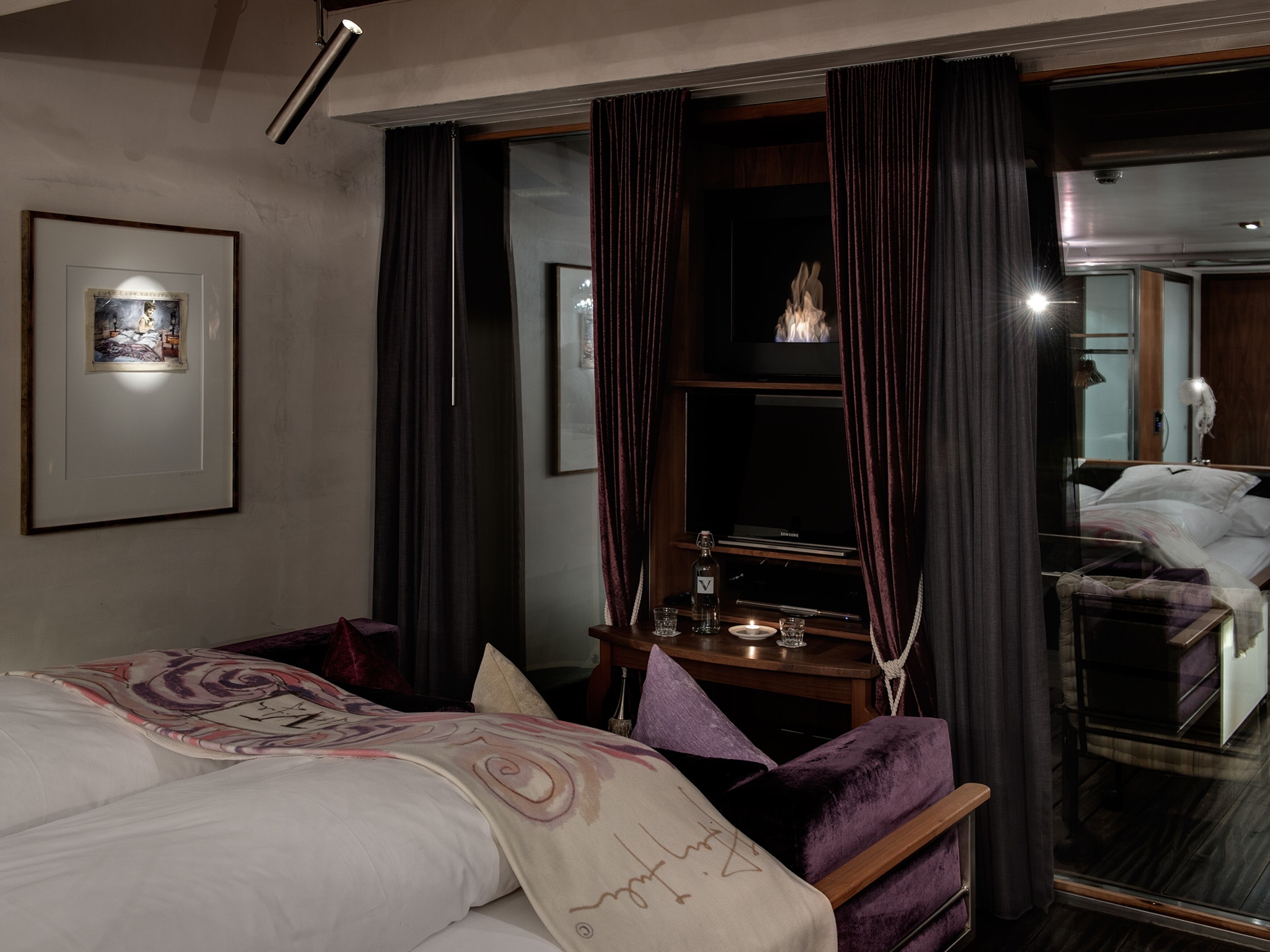 A hotel room with a bed, a desk with a lamp, a TV, and a mirror reflecting the room.