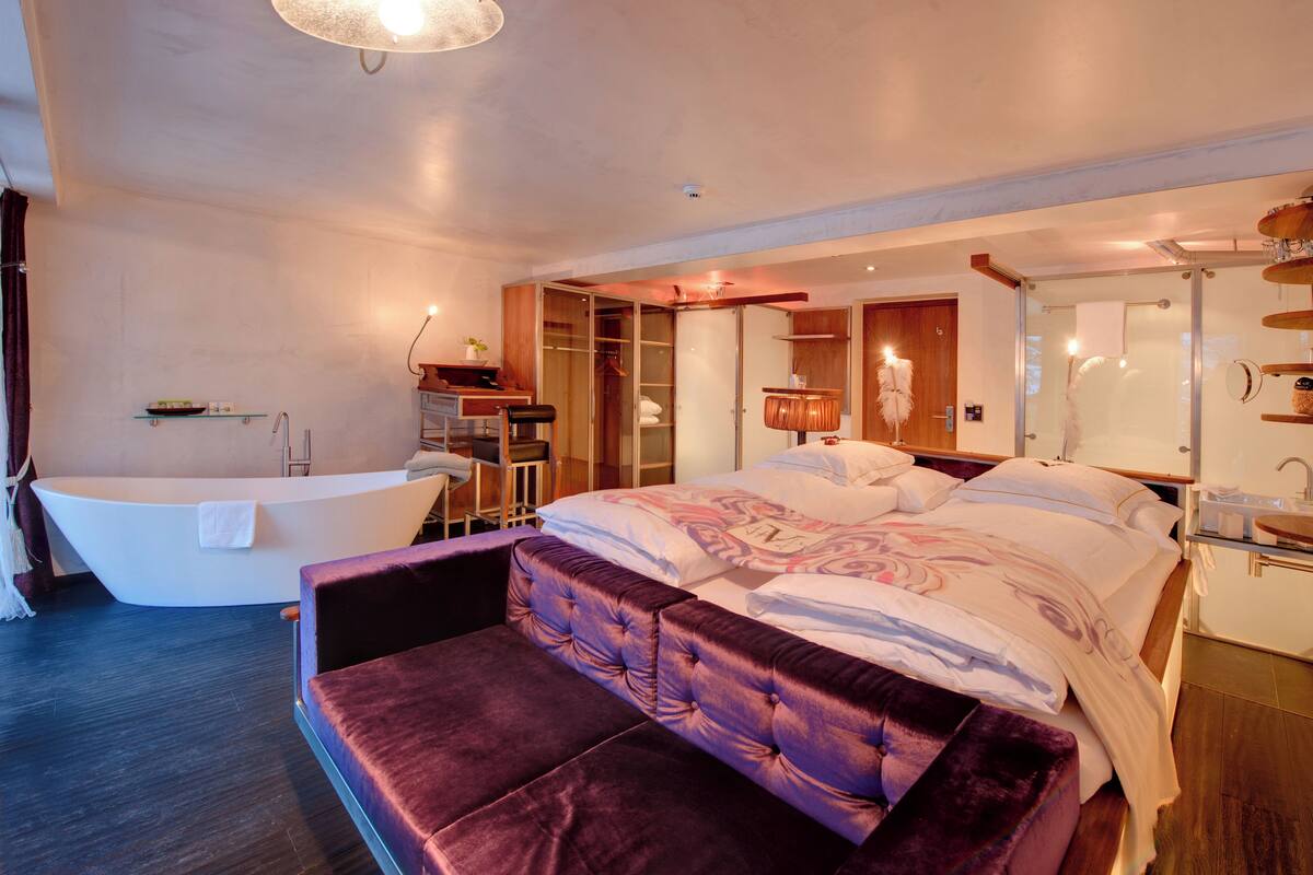 A modern bedroom with a large bed, a separate sitting area, a bathtub, and a walk-in closet.