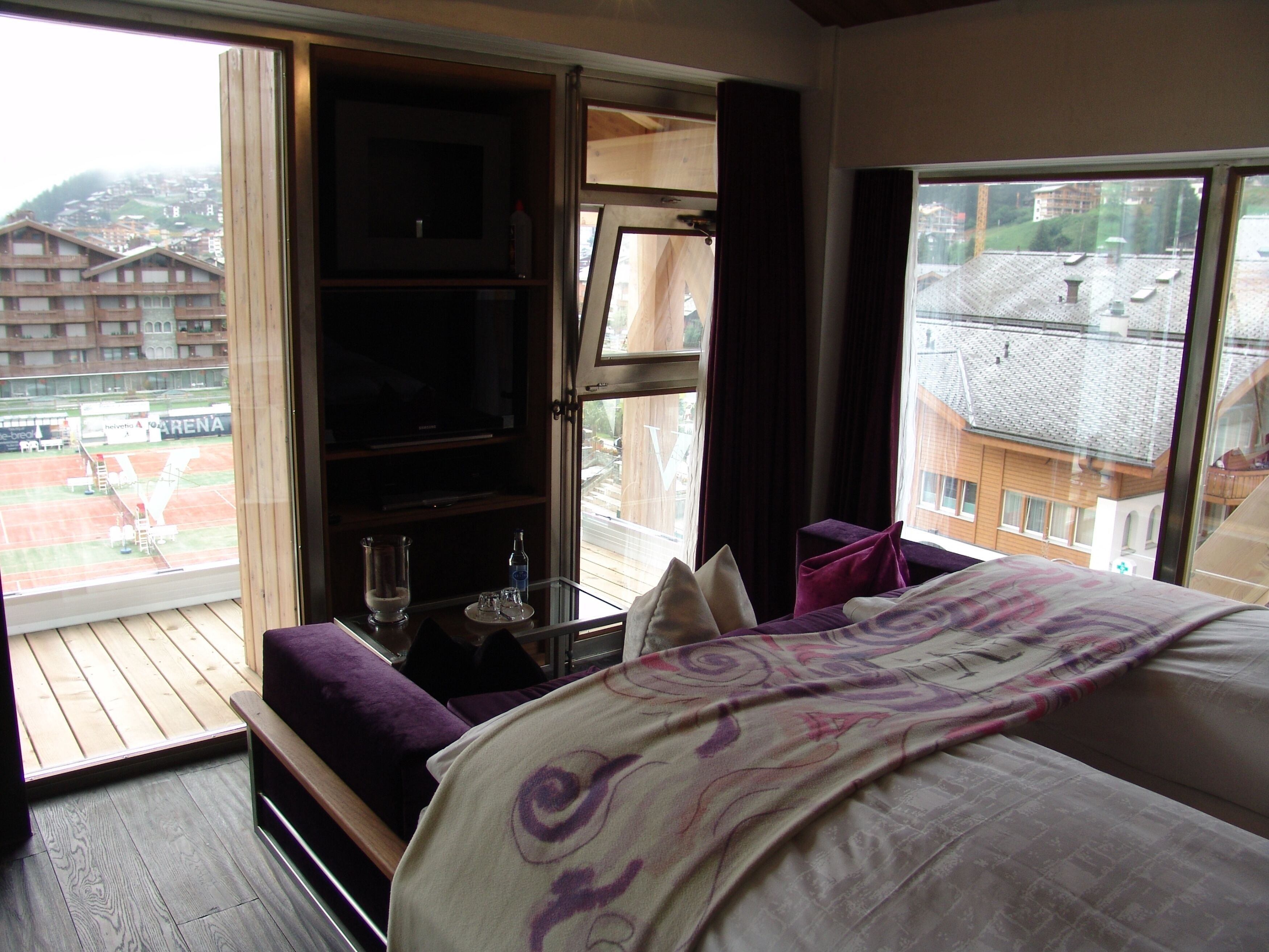 Habitación de hotel con cama, una mesita con vasos, estante con televisor y vista a un campo deportivo y edificios.