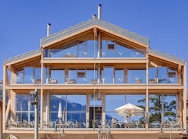 Un edificio de madera de varios pisos con grandes ventanales, mesas al aire libre y vistas a las montañas.